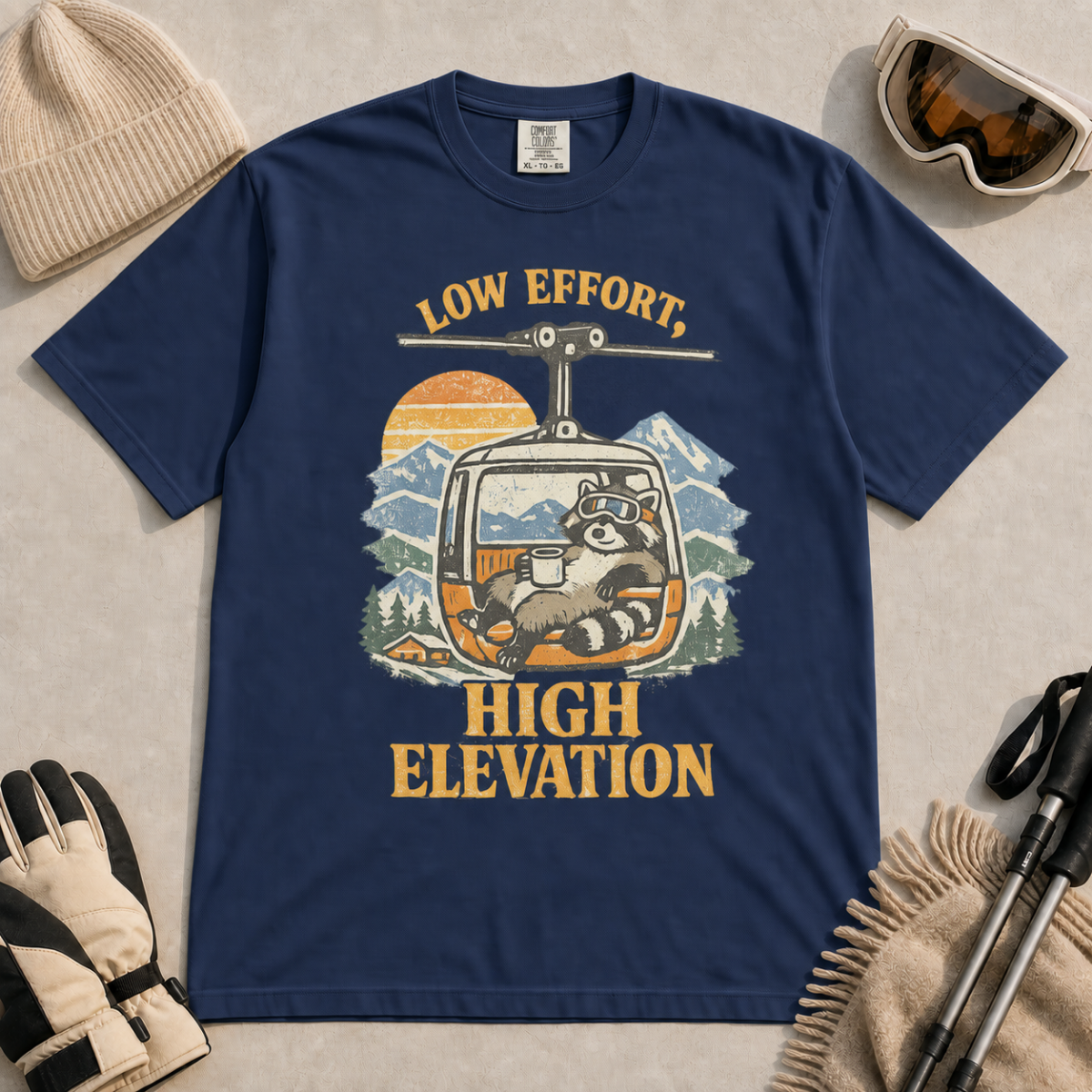 Navy t-shirt featuring a relaxed raccoon holding a mug inside a mountain gondola beneath the words “Low Effort, High Elevation.”