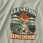 Close-up of a bay green t-shirt design featuring a small mountain rodent holding a mug beside ski goggles, with sunset peaks and the phrase “Elevation Without Expectation.”