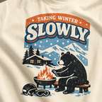 Close-up of a bear sitting beside a winter fire with snowy mountains, a cabin, helmet and goggles.