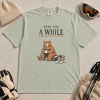 Bay green t-shirt featuring a seated bear holding a mug beside ski boots and goggles with the phrase “Here for a While.”