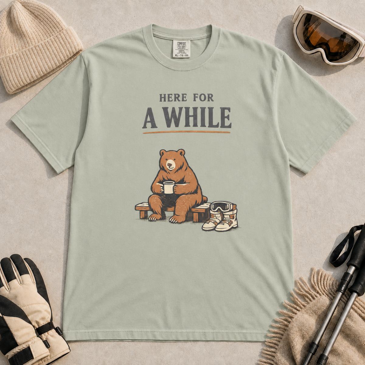 Bay green t-shirt featuring a seated bear holding a mug beside ski boots and goggles with the phrase “Here for a While.”