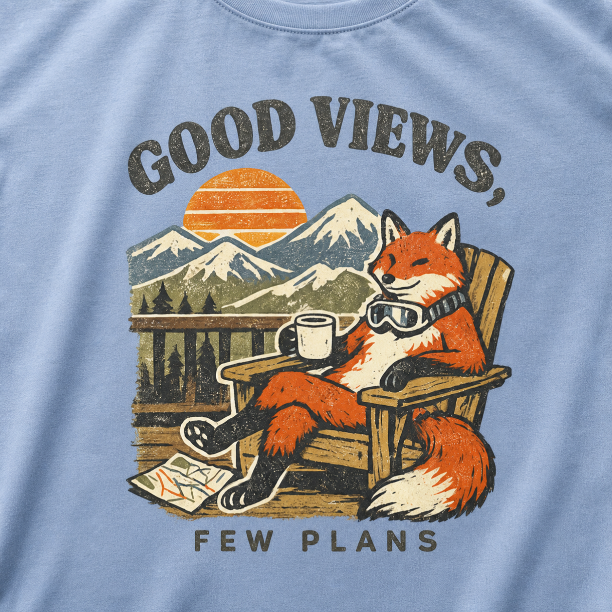 Close-up of a washed denim blue t-shirt design showing a fox relaxing with a mug before mountain scenery and the phrase “Good Views, Few Plans.”