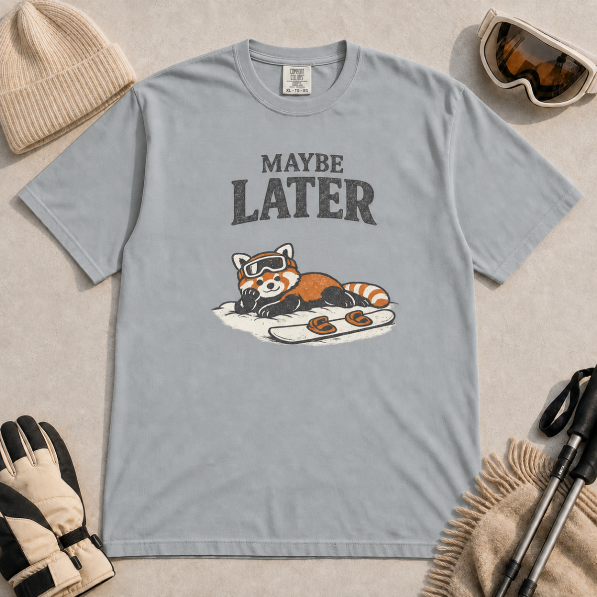 Granite gray t-shirt featuring a relaxed red panda in ski goggles lying beside a snowboard beneath the words “Maybe Later.”