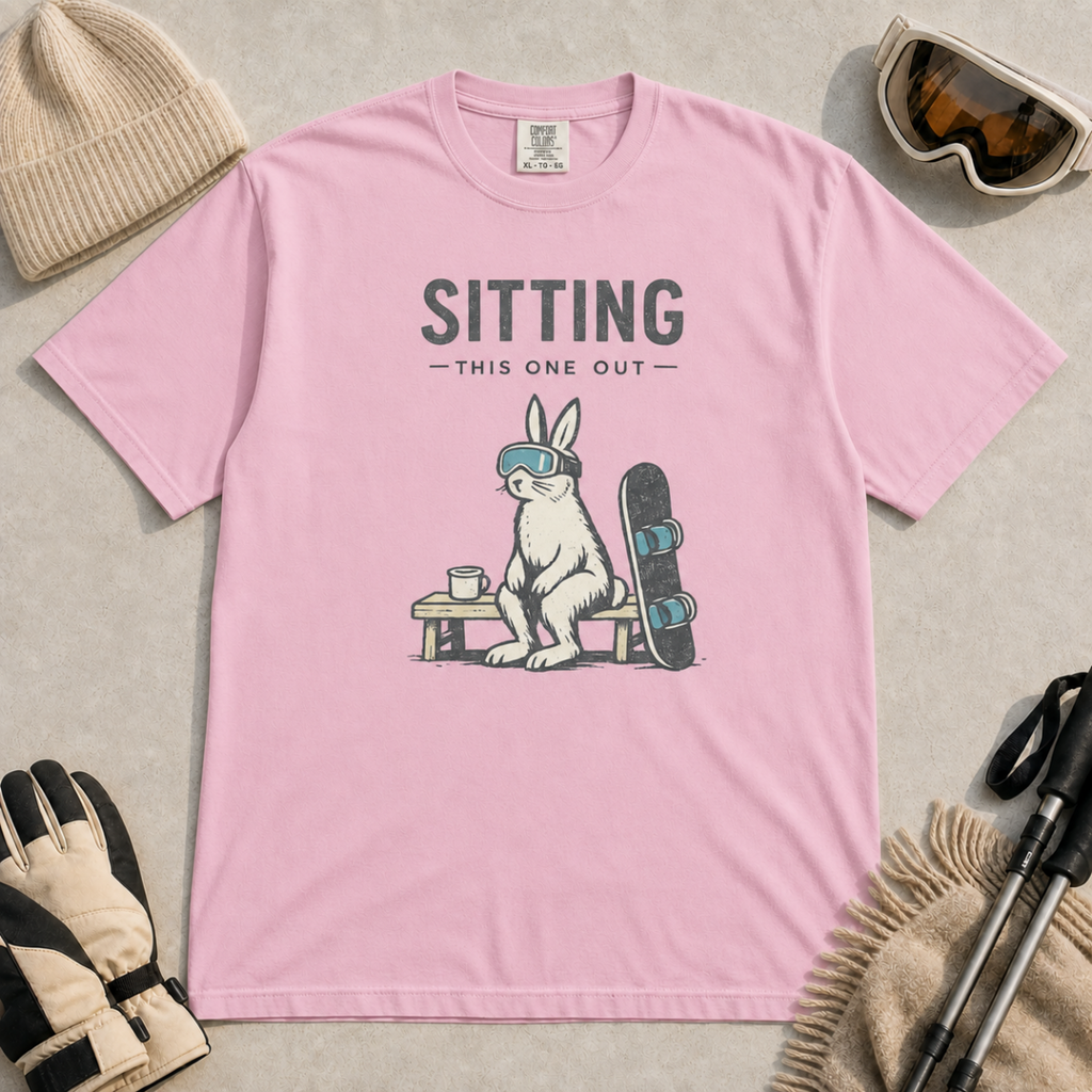 Blossom pink t-shirt featuring a goggle-wearing rabbit resting on a bench beside a mug and snowboard with “Sitting This One Out” text.