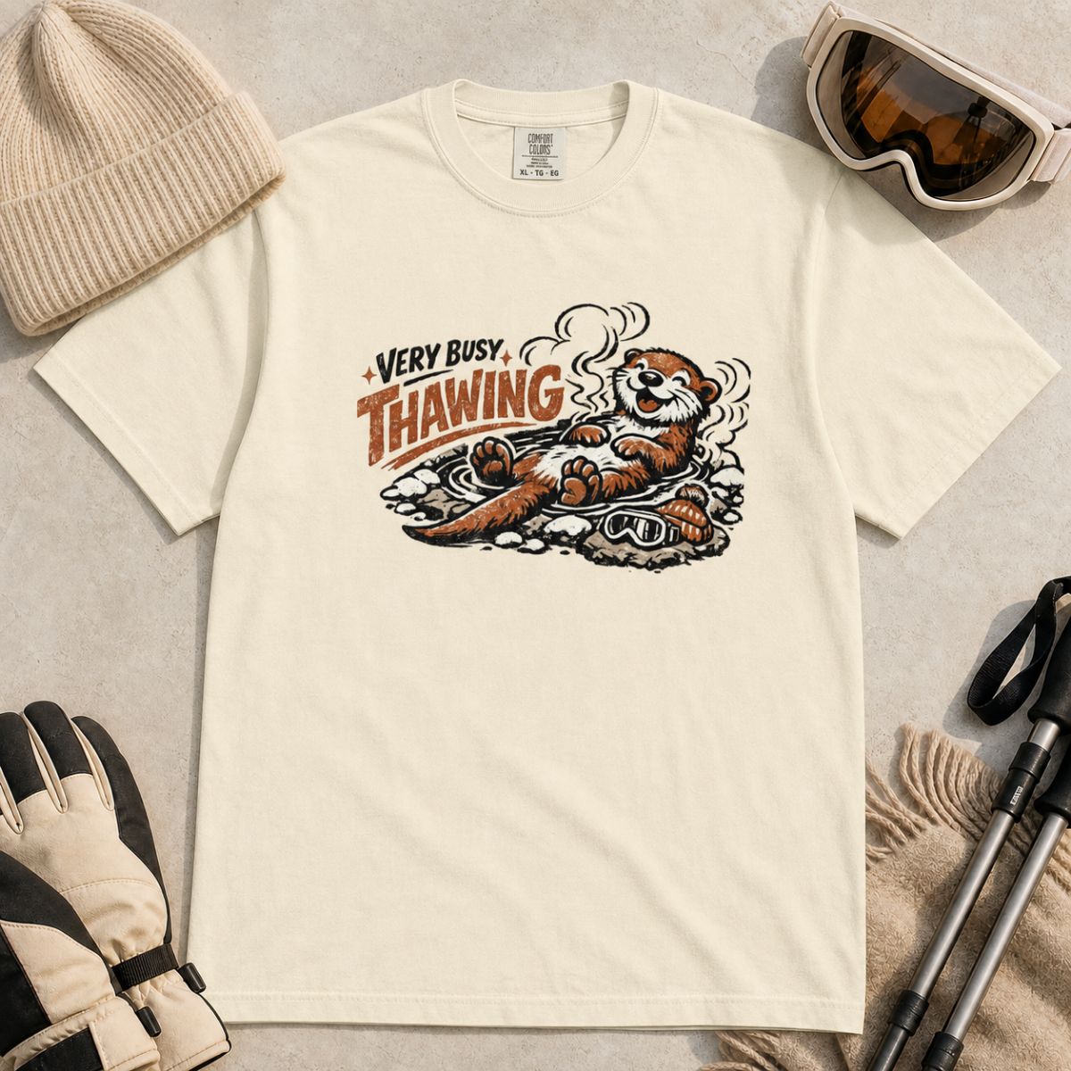 Ivory white t-shirt featuring a smiling otter thawing in a rocky hot spring beside ski goggles and a beanie.