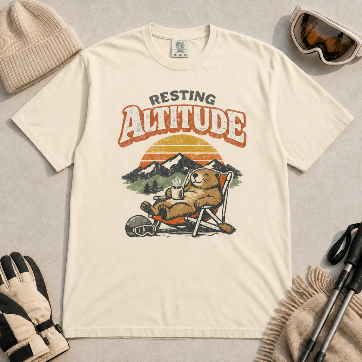 Ivory white t-shirt featuring a marmot resting in a chair with a mug, ski helmet and goggles beneath “Resting Altitude” text.