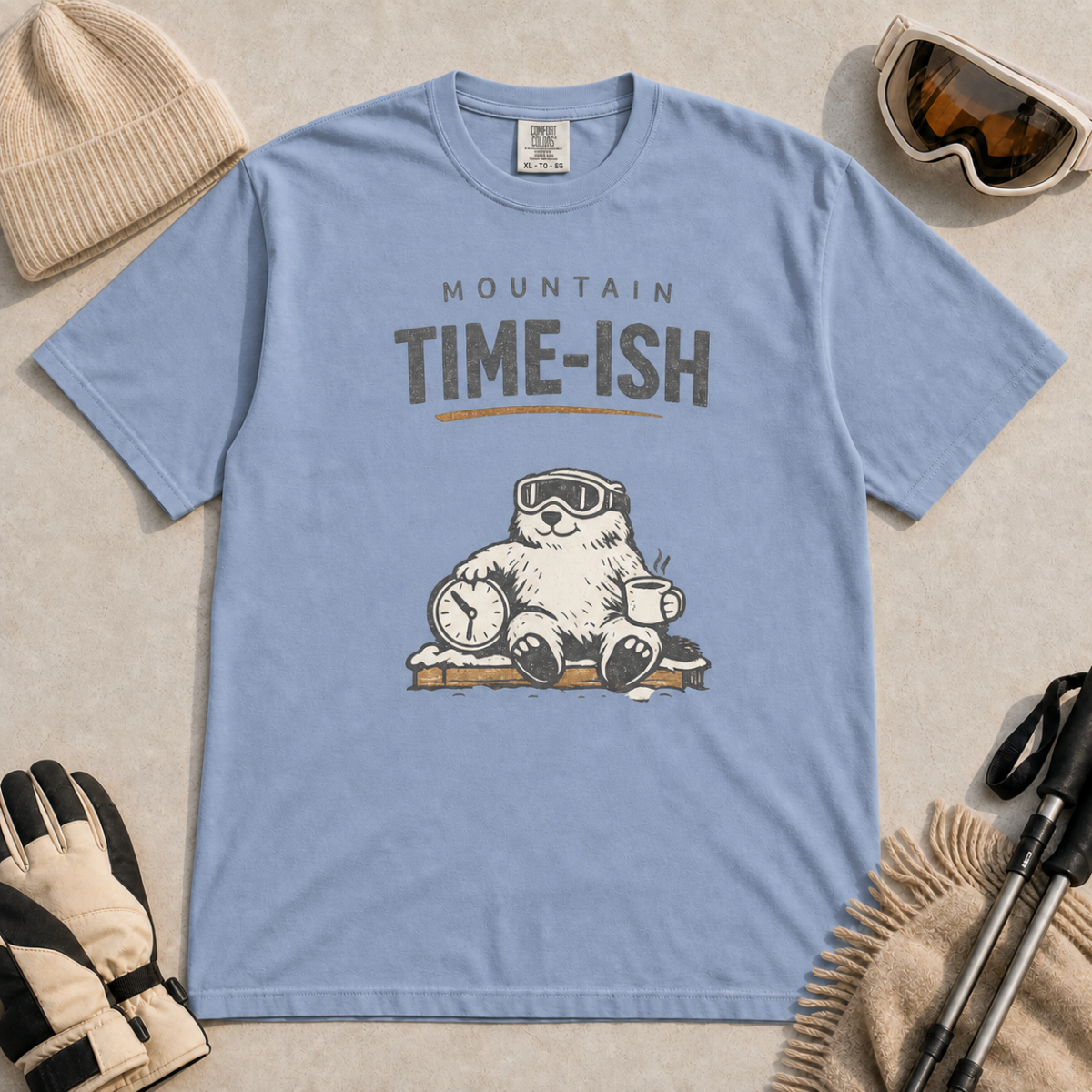 Washed denim blue t-shirt featuring a relaxed marmot in ski goggles holding a clock and steaming mug beneath the words “Mountain Time-ish.”