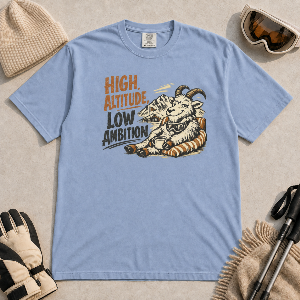 Washed denim blue t-shirt featuring a relaxed mountain goat holding a mug beside the phrase “High Altitude, Low Ambition.”