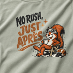 Close-up of a bay green t-shirt featuring a relaxed red squirrel in ski goggles reclining in a chair with a steaming mug, beside the words “No Rush, Just Après.”