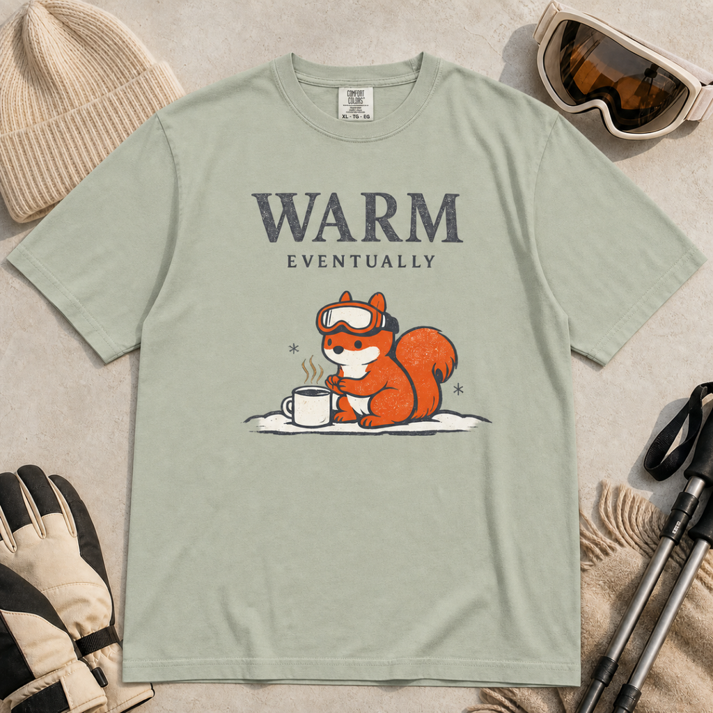 Bay green t-shirt featuring a bundled-up squirrel warming its paws beside a steaming mug under “Warm Eventually” text.
