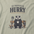 Close-up of a bay green t-shirt featuring a badger seated on a snowy bench with a mug, ski poles and goggles beneath the words “No Particular Hurry.”