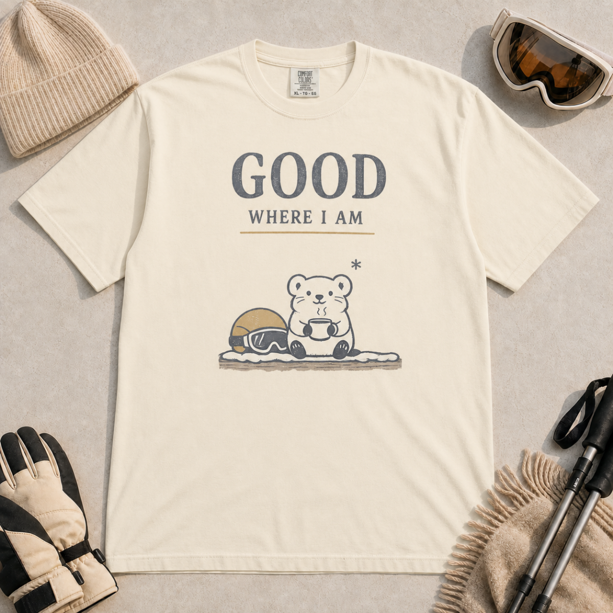 Ivory white t-shirt featuring a small white bear holding a mug beside ski goggles with the phrase “Good Where I Am.”