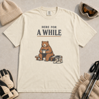 Ivory white t-shirt featuring a seated bear holding a mug beside ski boots and goggles with the phrase “Here for a While.”