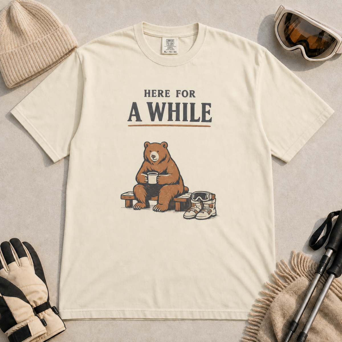 Ivory white t-shirt featuring a seated bear holding a mug beside ski boots and goggles with the phrase “Here for a While.”
