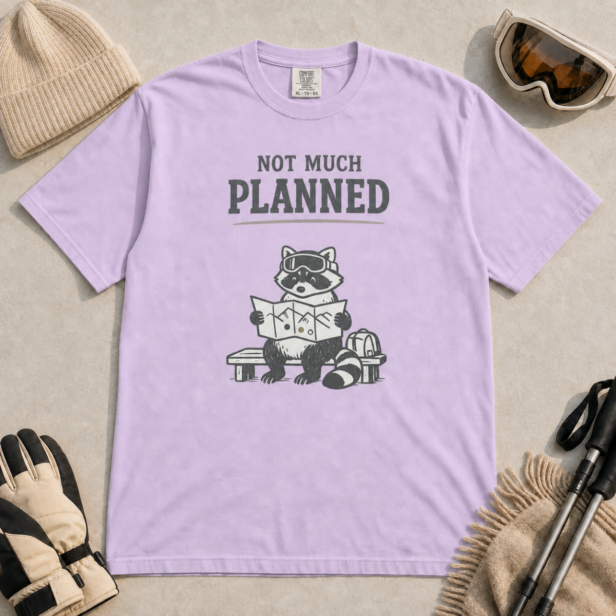 Orchid purple t-shirt featuring a raccoon in ski goggles sitting on a bench and studying a trail map, with the words “Not Much Planned.”