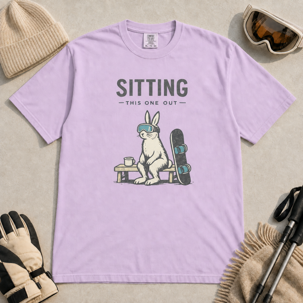 Orchid purple t-shirt featuring a goggle-wearing rabbit resting on a bench beside a mug and snowboard with “Sitting This One Out” text.