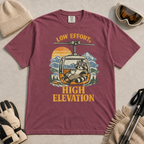 Brick red t-shirt featuring a relaxed raccoon holding a mug inside a mountain gondola beneath the words “Low Effort, High Elevation.”