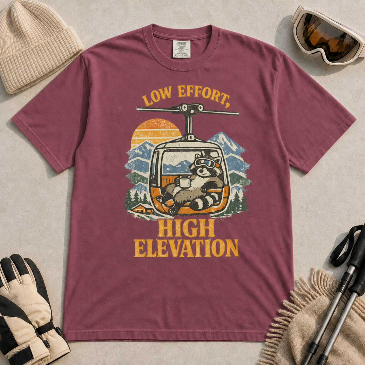 Brick red t-shirt featuring a relaxed raccoon holding a mug inside a mountain gondola beneath the words “Low Effort, High Elevation.”