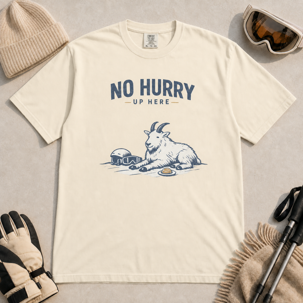 Ivory white t-shirt featuring a mountain goat relaxing in the snow with a helmet, goggles and snack beneath the words “No Hurry Up Here.”