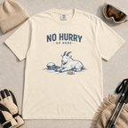 Ivory white t-shirt featuring a mountain goat relaxing in the snow with a helmet, goggles and snack beneath the words “No Hurry Up Here.”