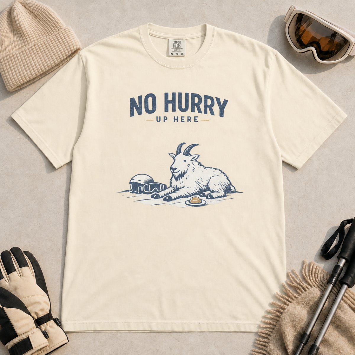 Ivory white t-shirt featuring a mountain goat relaxing in the snow with a helmet, goggles and snack beneath the words “No Hurry Up Here.”