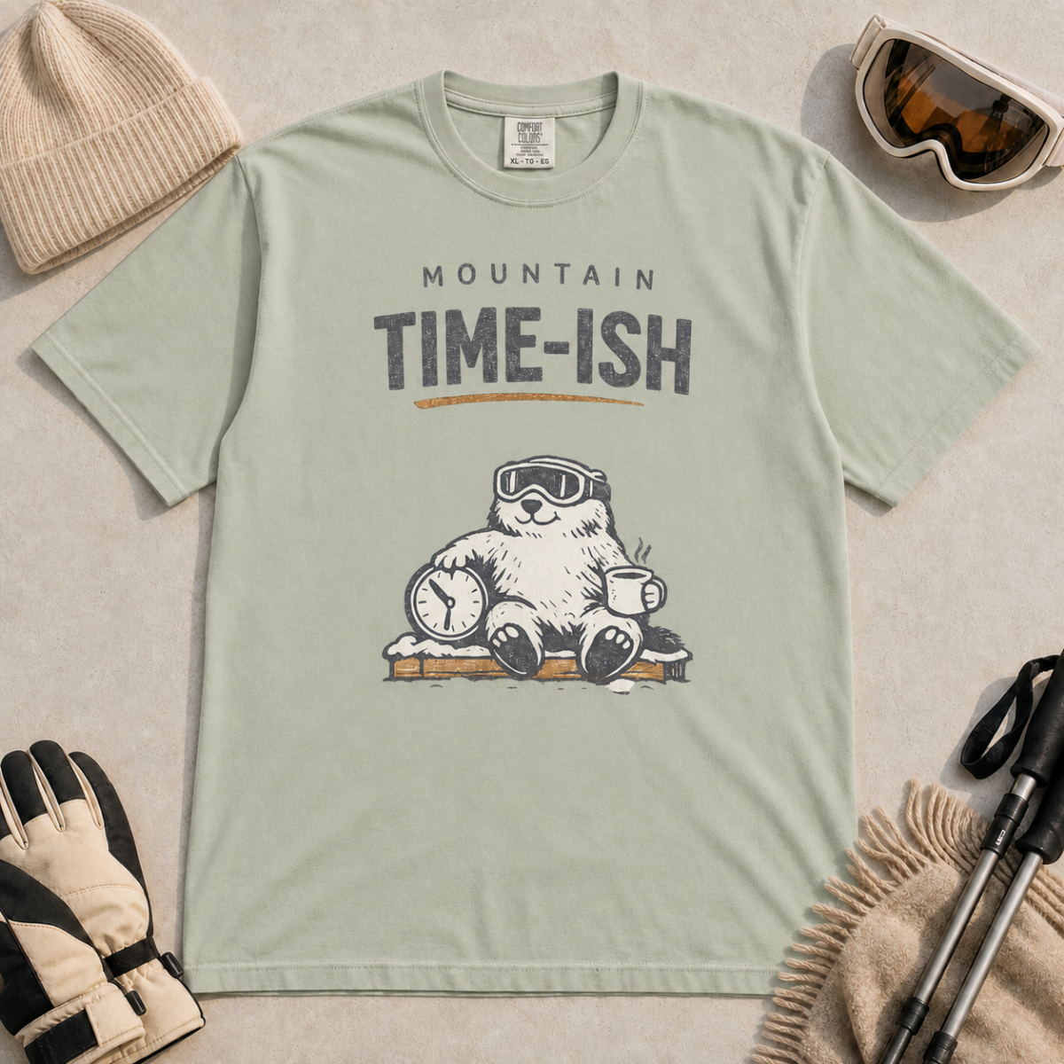 Bay green t-shirt featuring a relaxed marmot in ski goggles holding a clock and steaming mug beneath the words “Mountain Time-ish.”