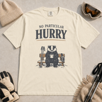 Ivory white t-shirt featuring a badger relaxing on a snowy bench with a mug, ski poles and goggles beneath the words “No Particular Hurry.”