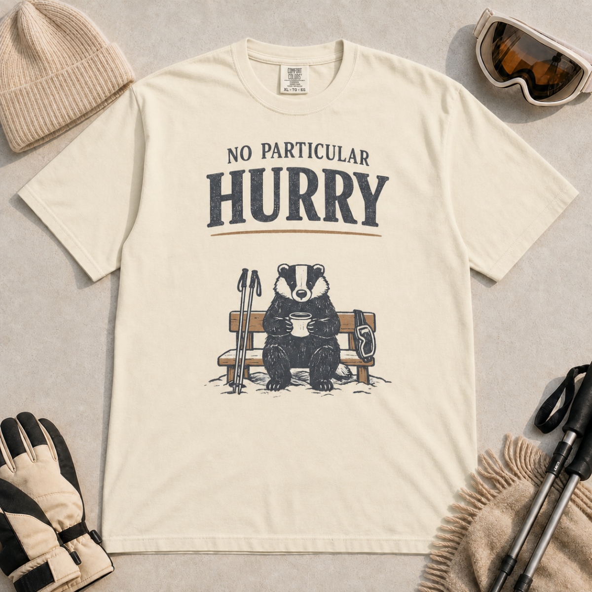 Ivory white t-shirt featuring a badger relaxing on a snowy bench with a mug, ski poles and goggles beneath the words “No Particular Hurry.”