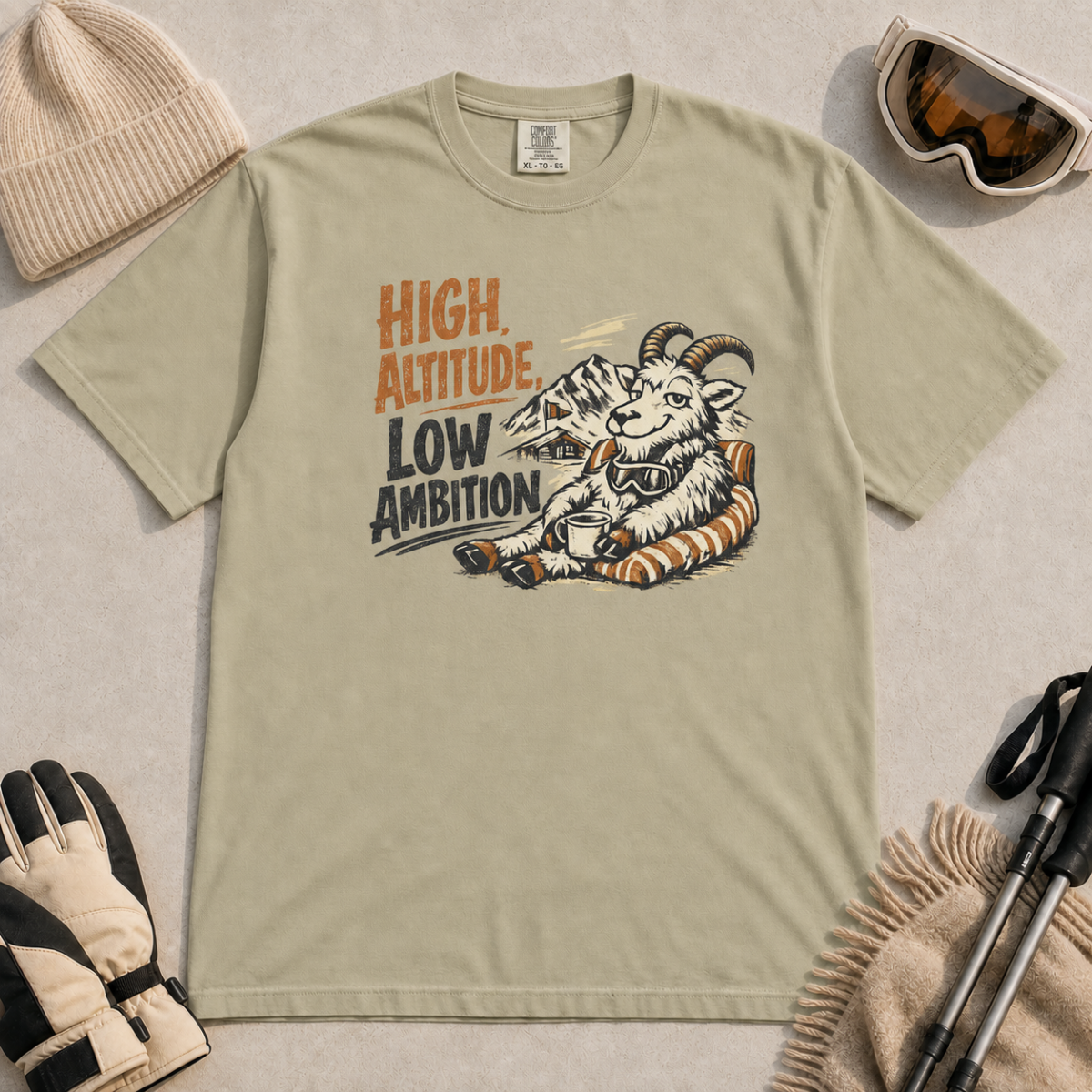 Khaki t-shirt featuring a relaxed mountain goat holding a mug beside the phrase “High Altitude, Low Ambition.”