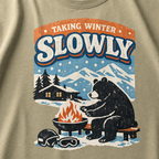 Close-up of a bear sitting beside a winter fire with snowy mountains, a cabin, helmet and goggles.
