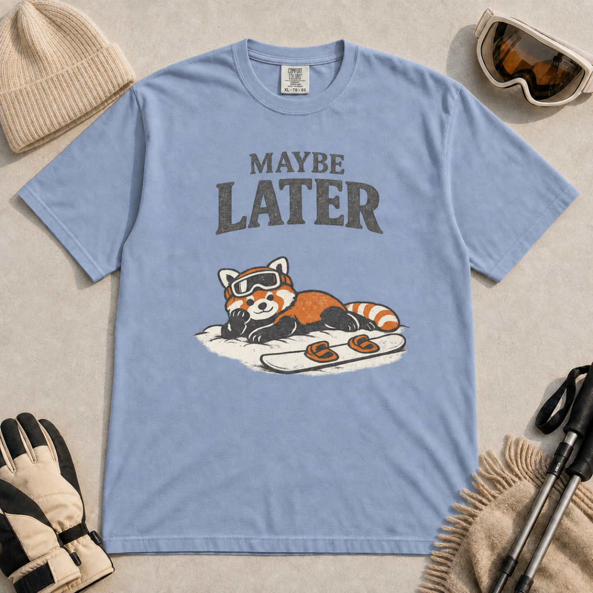 Washed denim blue t-shirt featuring a relaxed red panda in ski goggles lying beside a snowboard beneath the words “Maybe Later.”