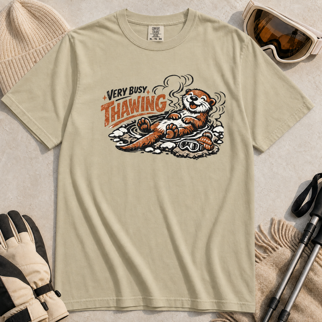 Khaki t-shirt featuring a smiling otter thawing in a rocky hot spring beside ski goggles and a beanie.