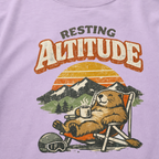 Close-up of a relaxed marmot reclining in a chair with a steaming mug beneath “Resting Altitude” lettering and a mountain sunset.
