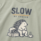 Close-up of a seated hedgehog resting beside ski goggles and a steaming mug under “Slow by Choice” lettering.