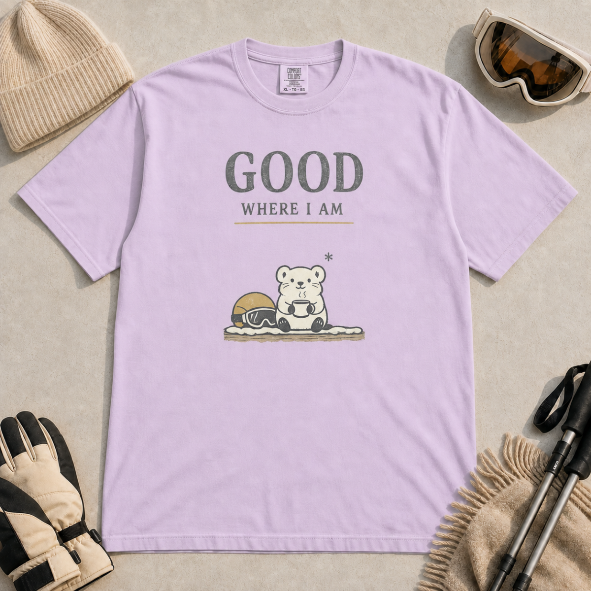 Orchid purple t-shirt featuring a small white bear holding a mug beside ski goggles with the phrase “Good Where I Am.”
