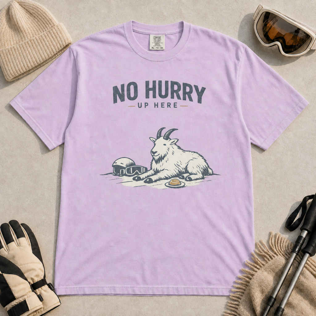 Orchid purple t-shirt featuring a mountain goat relaxing in the snow with a helmet, goggles and snack beneath the words “No Hurry Up Here.”