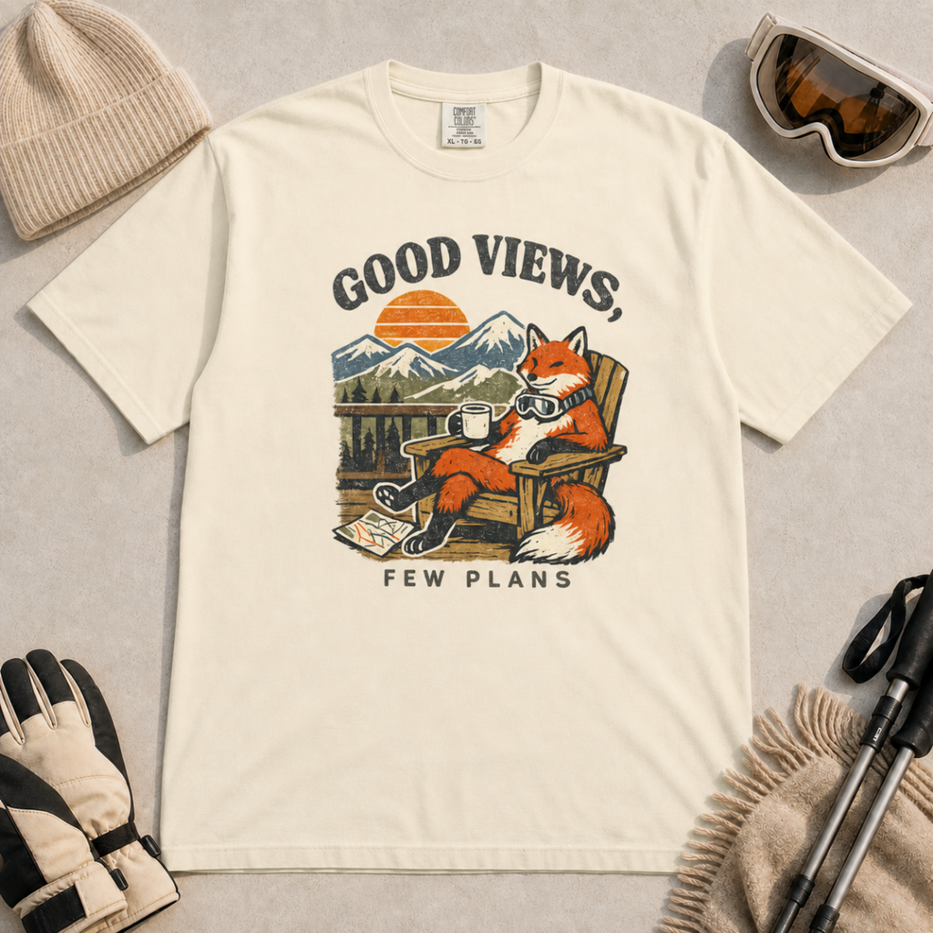 Ivory white t-shirt featuring a relaxed fox in a wooden chair holding a mug against a mountain sunset with the phrase “Good Views, Few Plans.”