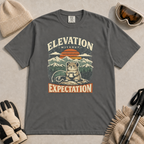 Pepper grey t-shirt featuring a mountain rodent with a mug and ski goggles beneath retro sunset scenery and the phrase “Elevation Without Expectation.”