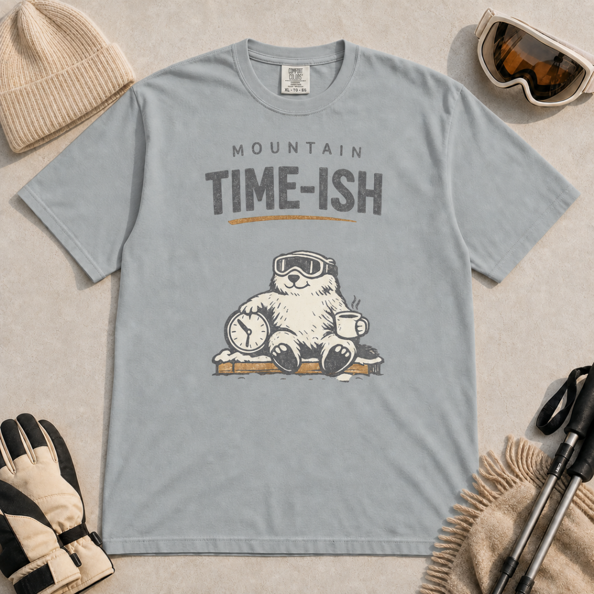 Granite gray t-shirt featuring a relaxed marmot in ski goggles holding a clock and steaming mug beneath the words “Mountain Time-ish.”