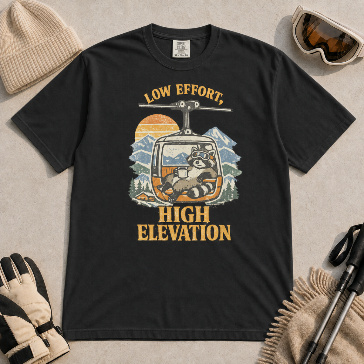 Black t-shirt featuring a relaxed raccoon holding a mug inside a mountain gondola beneath the words “Low Effort, High Elevation.”
