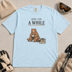 Chambray blue t-shirt featuring a seated bear holding a mug beside ski boots and goggles with the phrase “Here for a While.”