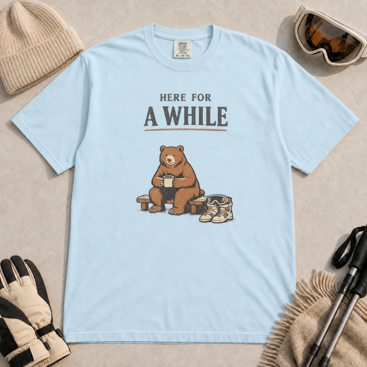 Chambray blue t-shirt featuring a seated bear holding a mug beside ski boots and goggles with the phrase “Here for a While.”