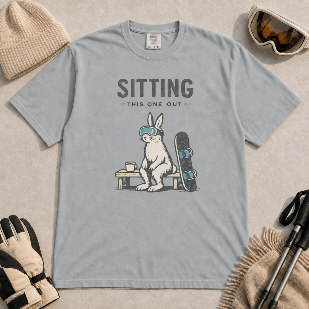 Granite gray t-shirt featuring a goggle-wearing rabbit resting on a bench beside a mug and snowboard with “Sitting This One Out” text.