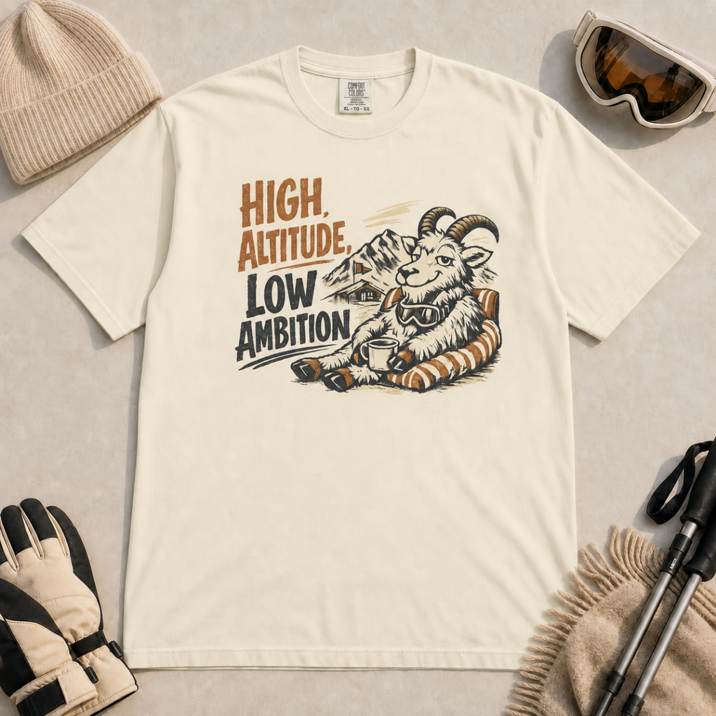 Ivory white t-shirt featuring a relaxed mountain goat holding a mug beside the phrase “High Altitude, Low Ambition.”