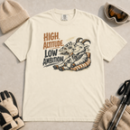 Ivory white t-shirt featuring a relaxed mountain goat holding a mug beside the phrase “High Altitude, Low Ambition.”