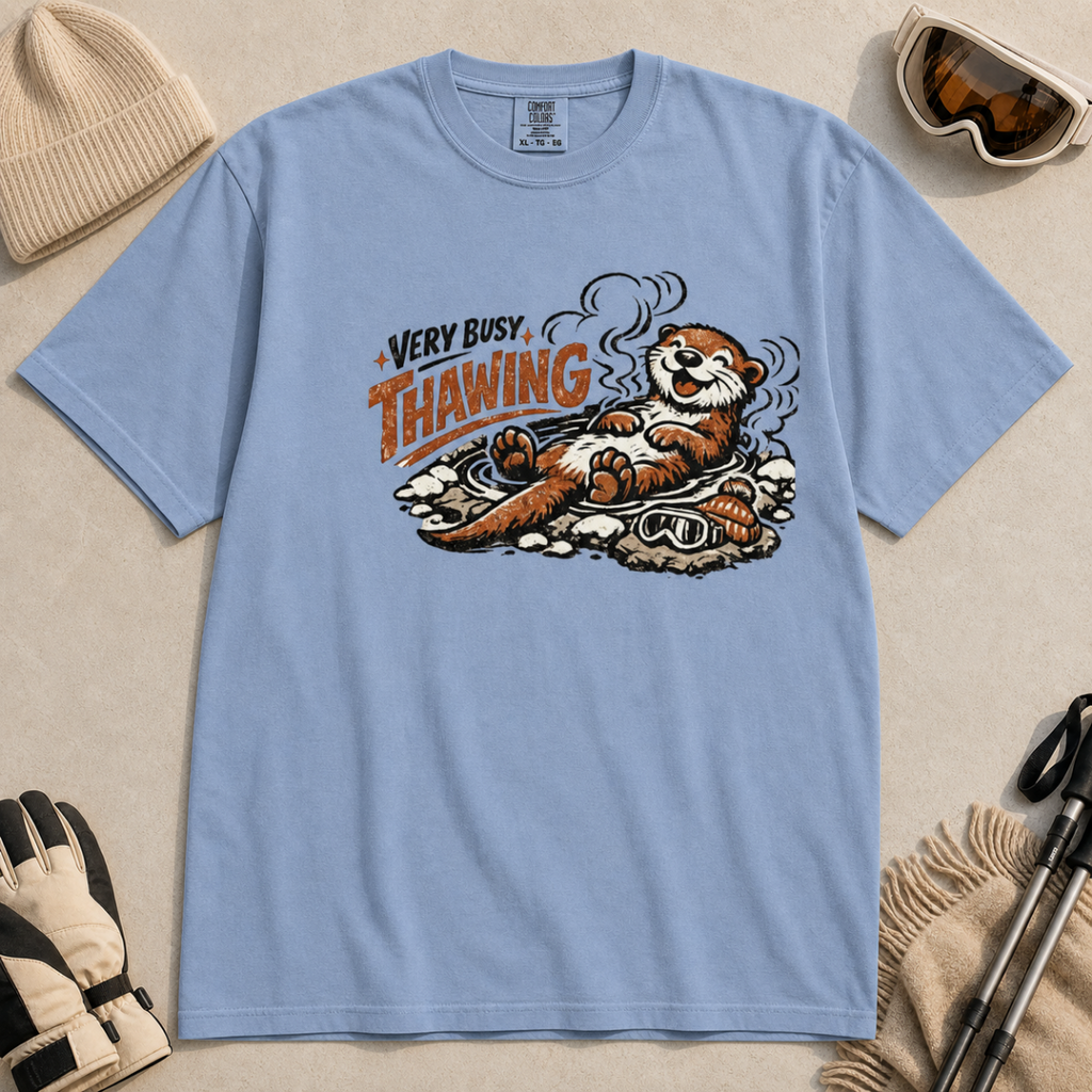 Washed denim blue t-shirt featuring a smiling otter thawing in a rocky hot spring beside ski goggles and a beanie.