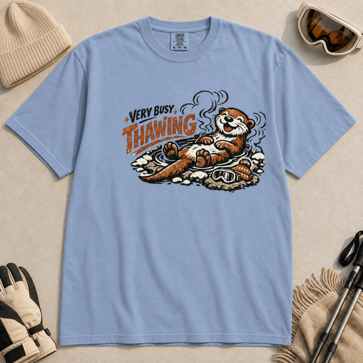 Washed denim blue t-shirt featuring a smiling otter thawing in a rocky hot spring beside ski goggles and a beanie.