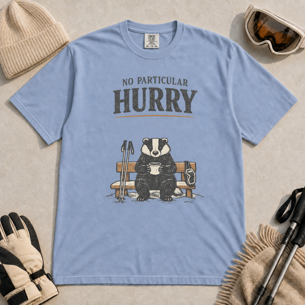 Washed denim blue t-shirt featuring a badger relaxing on a snowy bench with a mug, ski poles and goggles beneath the words “No Particular Hurry.”