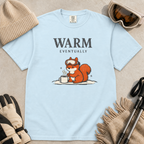 Chambray blue t-shirt featuring a bundled-up squirrel warming its paws beside a steaming mug under “Warm Eventually” text.