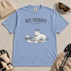 Washed denim blue t-shirt featuring a mountain goat relaxing in the snow with a helmet, goggles and snack beneath the words “No Hurry Up Here.”